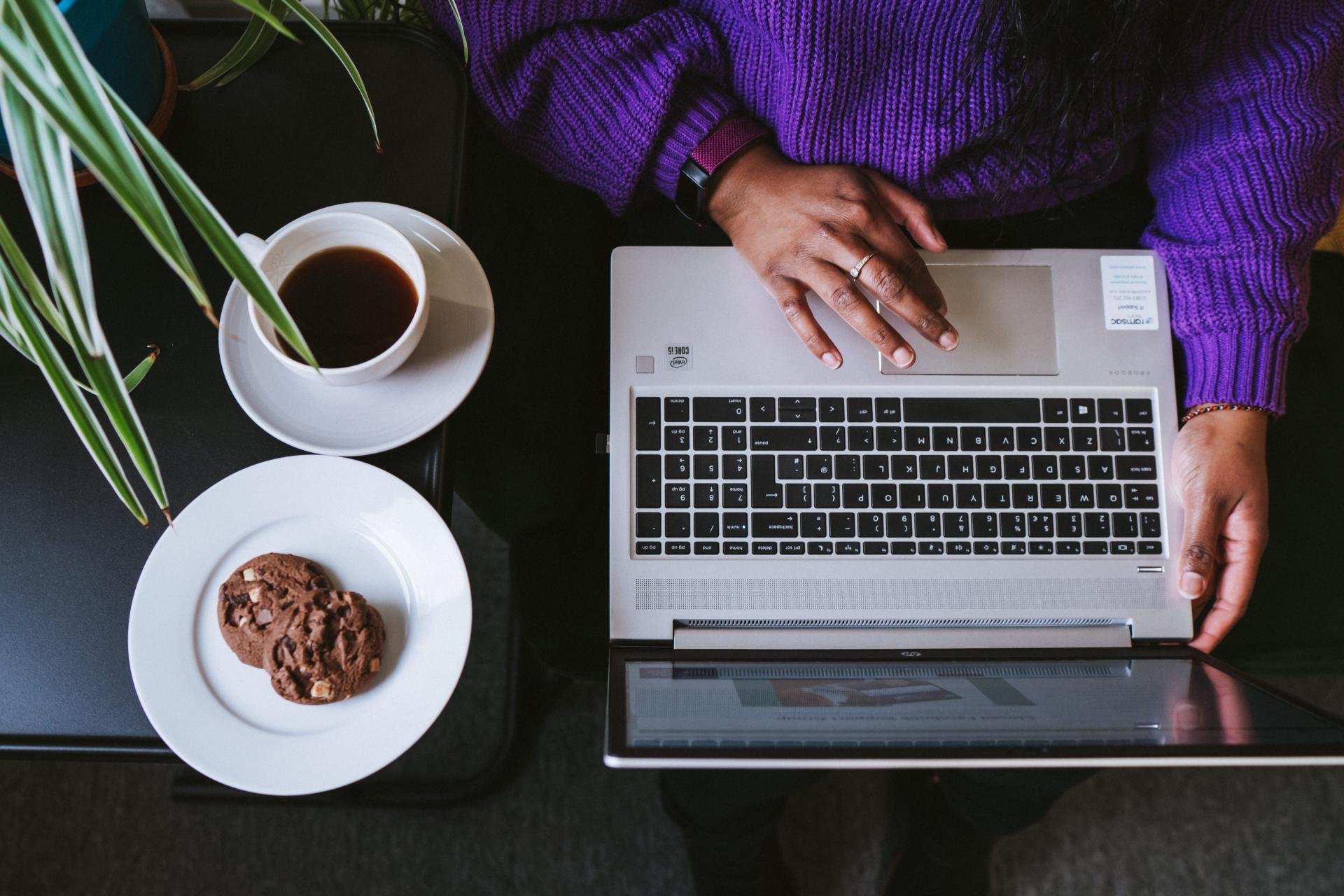 Top down view of woman on laptop with a cup of tea and some biscuits next to her.