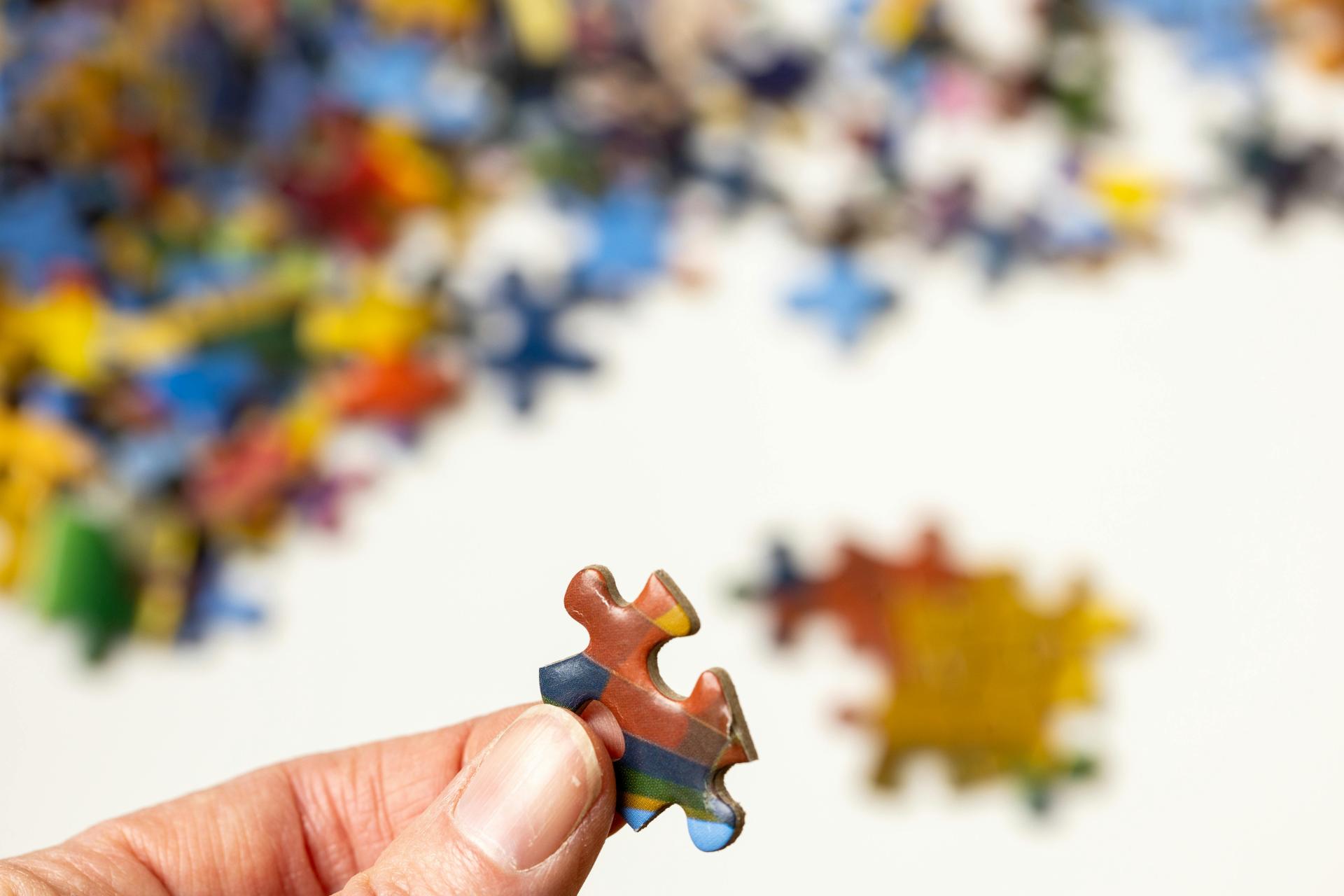 A hand is holding a jigsaw puzzle piece between thumb and finger, with other pieces in the background