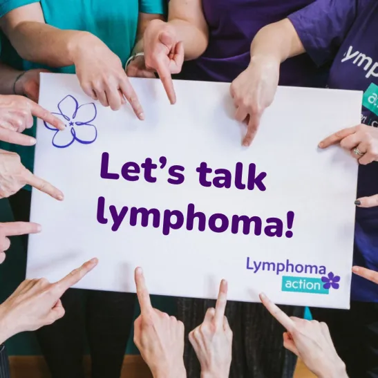 A close up of a sign with text 'let's talk lymphoma!'. A group of people surround the sign, with the focus on their hands pointing to the text.