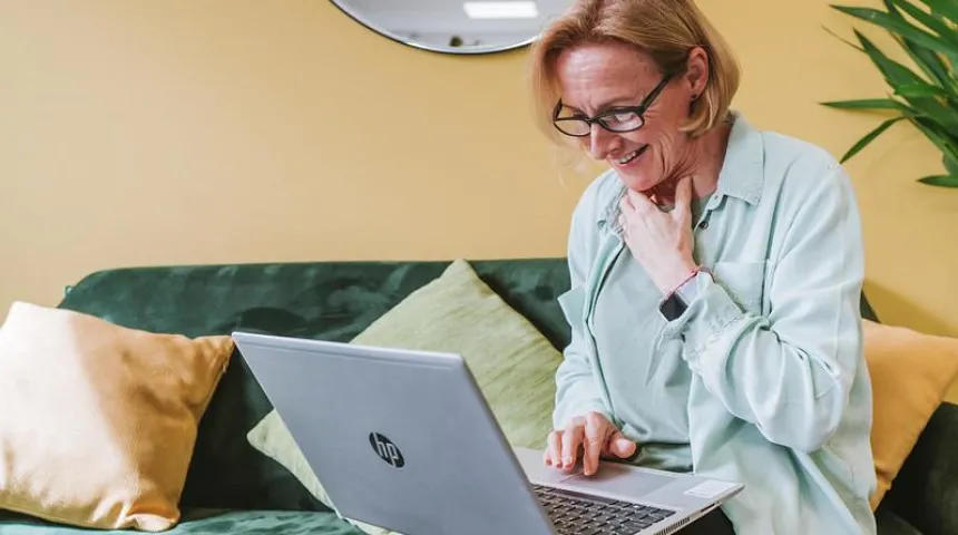 A person, smiling, sits on a sofa and looks at a laptop screen.