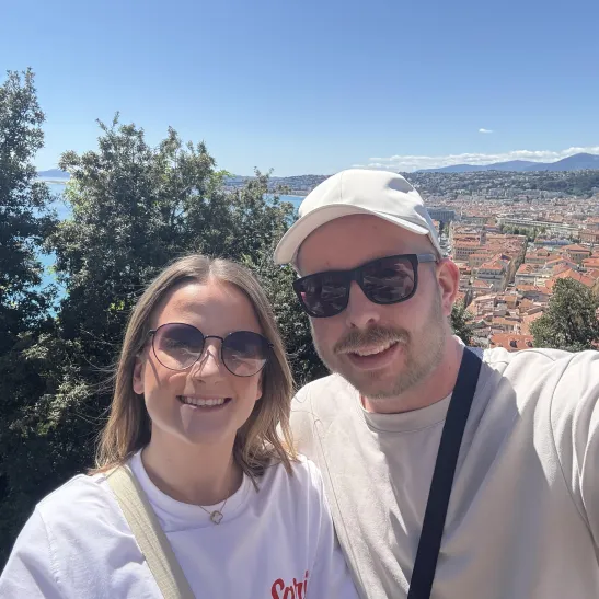 Bradley stands to the right with his arm around his partner, Mia. Both are smiling to the camera against a scenic backdrop of trees, an ocean view and coast side houses. Mountains can be seen in the far distance. 