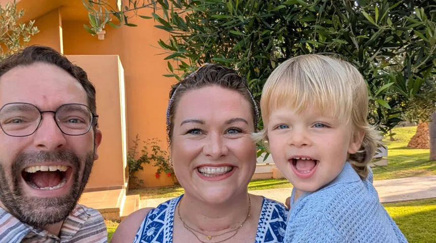 Rebecca is centre and is holding her son. Her husband is to her left. All three are smiling to camera. A tree green leaves is visible in the background.