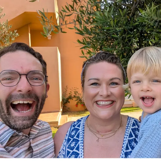 Rebecca is centre and is holding her son. Her husband is to her left. All three are smiling to camera. A tree green leaves is visible in the background.