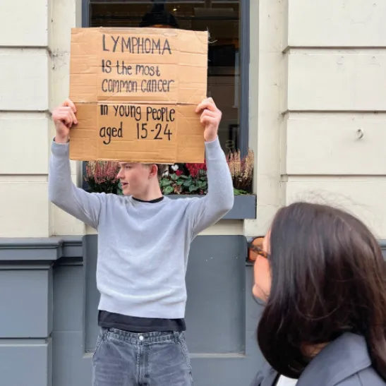 An image of a young person holding up a sign that reads: 'Lymphoma is the most common cancer in young people aged 15-24'